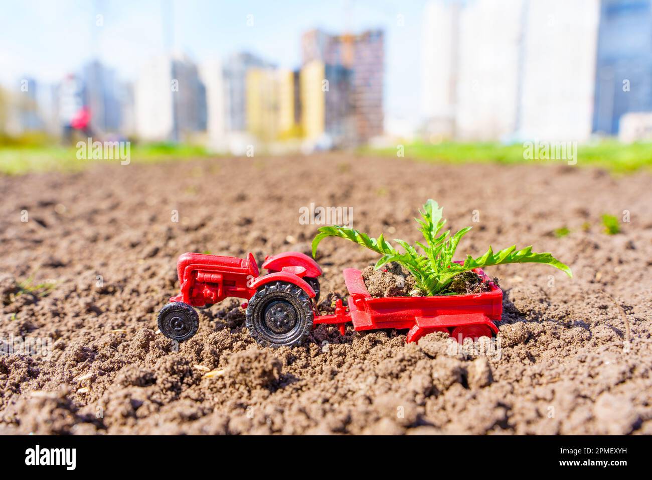 Agriculture vs urbanisation : tracteur jouet rouge avec une remorque remplie de jeunes pousses vertes debout sur des terres fraîchement labourées, avec une image floue d'une ville Banque D'Images