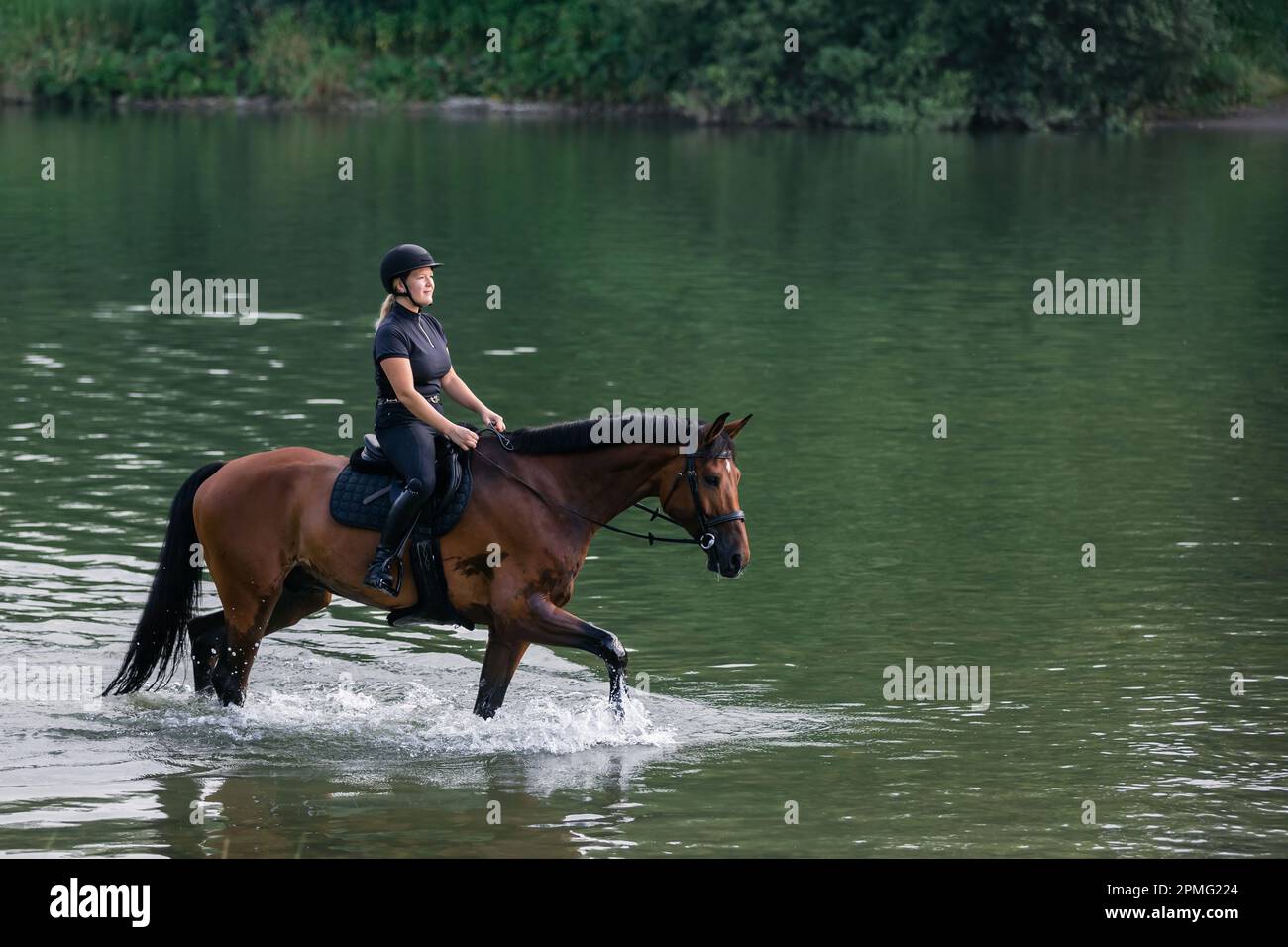 Une femelle à cheval dans un ensemble de jockey noir à cheval sur un cheval de châtaignier le long de la rivière au coucher du soleil. Concept de loisirs, d'équitation et de nature. Banque D'Images