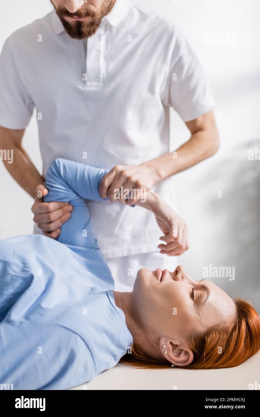 kinésithérapeute barbu fléchissant bras douloureux de la femme pendant la thérapie de réadaptation en clinique, image de stock Banque D'Images