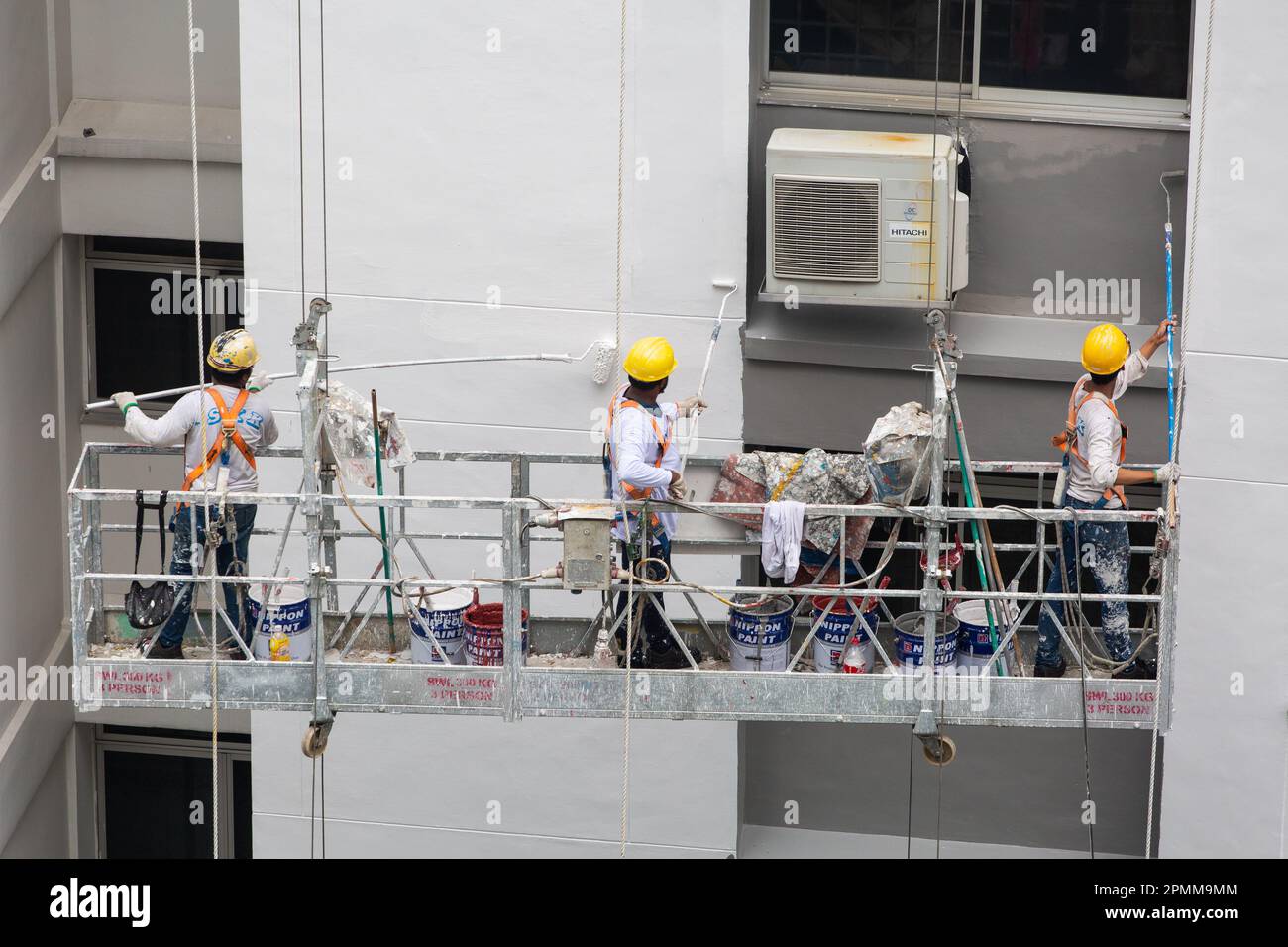 Trois travailleurs avec un casque de sécurité sur l'ascenseur de l'échafaudage peignant de la peinture fraîche sur l'extérieur de l'architecture. Banque D'Images
