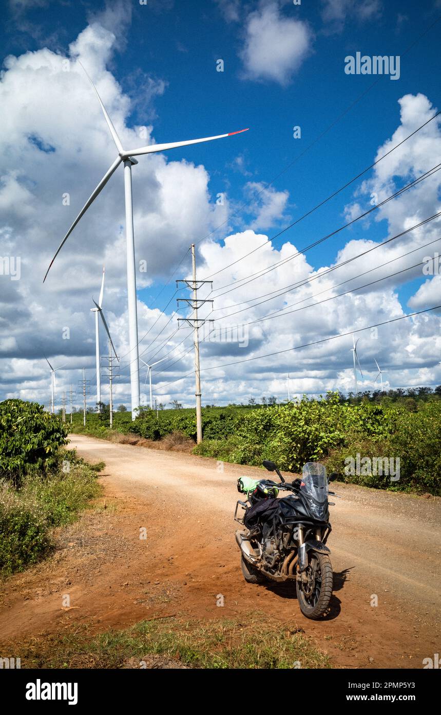 Une grosse moto noire garée sur une piste de terre à côté d'une ligne de transmission de puissance, d'énormes éoliennes et une plantation de café dans la province de Gia Lai Banque D'Images