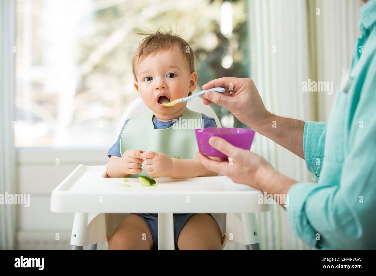 Père nourrissant son bébé mignon avec la première nourriture solide, bébé assis dans une chaise haute. Enfant goûtant des légumes à table, découvrant de nouveaux aliments. Banque D'Images