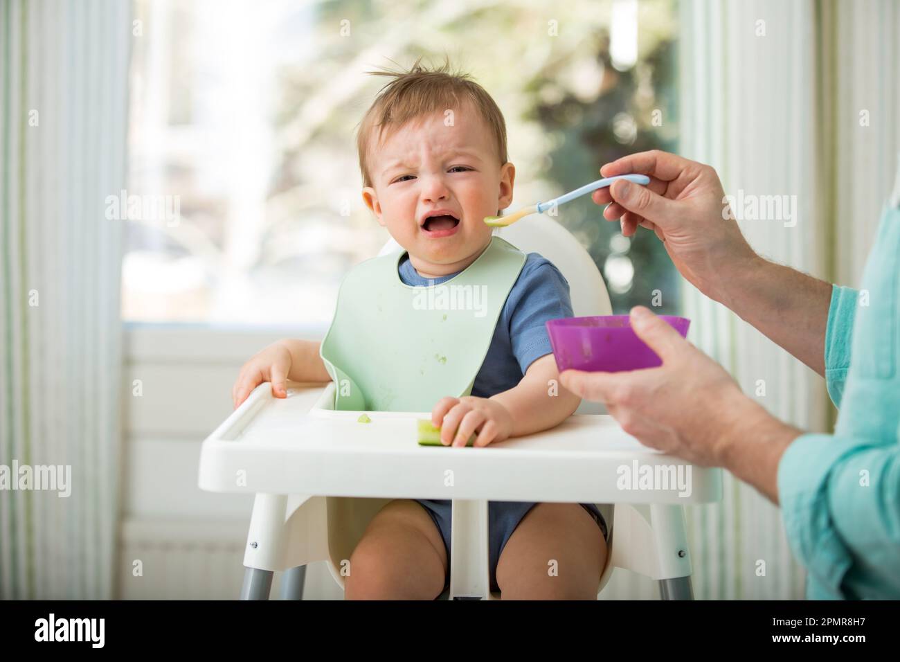 Père nourrissant son bébé mignon avec la première nourriture solide, bébé assis dans une chaise haute. L'enfant ne veut pas manger, refuse de manger, fait des visages. Cuisine confortable Banque D'Images