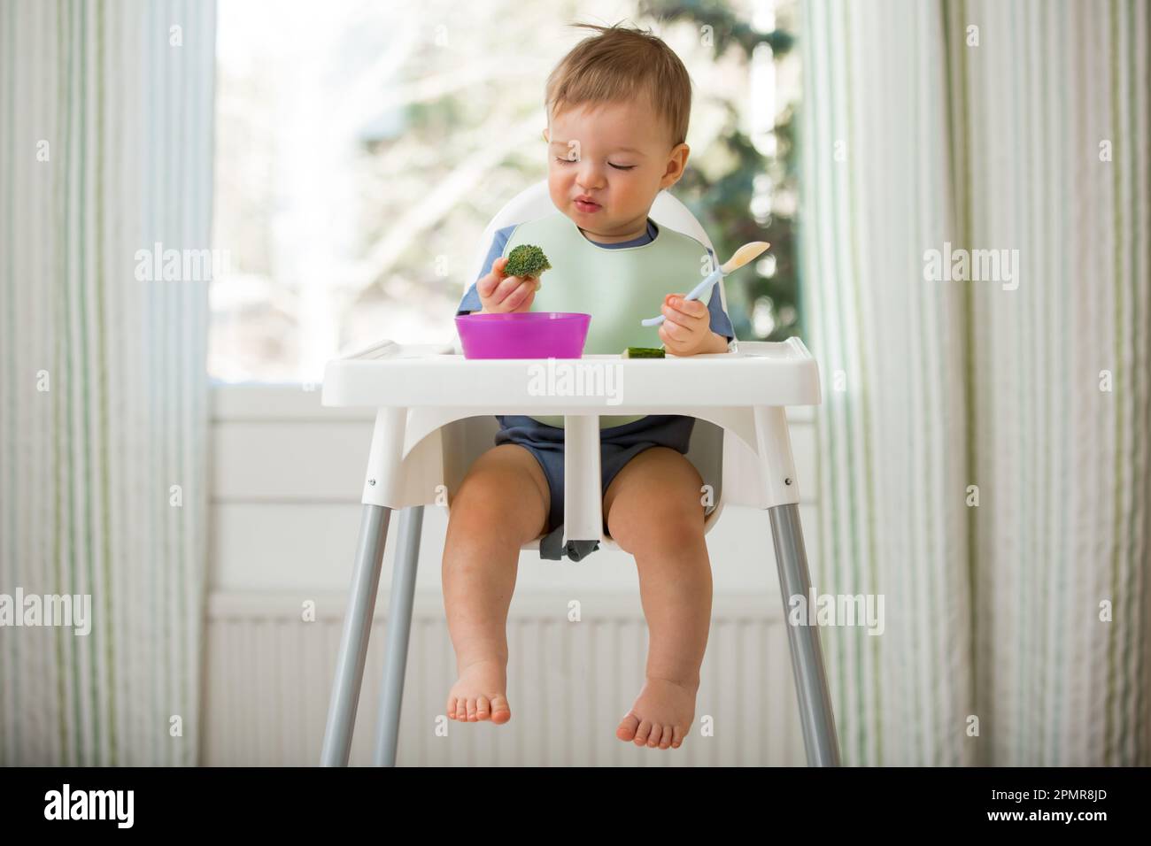 Bébé mignon mangeant avec la première nourriture solide, bébé assis dans une chaise haute. L'enfant ne veut pas manger, refuse de manger, se fait des visages. Intérieur de cuisine confortable. Banque D'Images