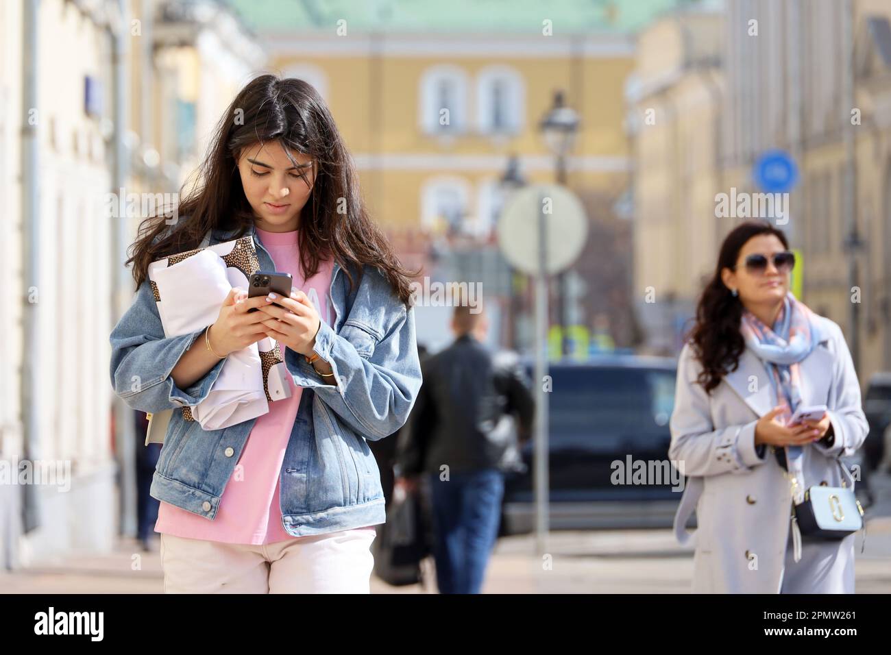 Les gens qui utilisent des téléphones mobiles, jolie fille en Jean veste marchant en premier plan avec smartphone dans les mains sur la rue de ville au printemps Banque D'Images