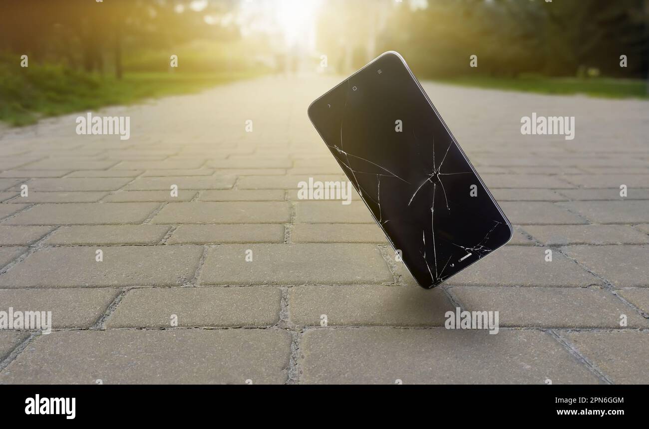 Un smartphone dont l'écran est cassé tombe sur un pont en face d'un parc et de la lumière du soleil Banque D'Images