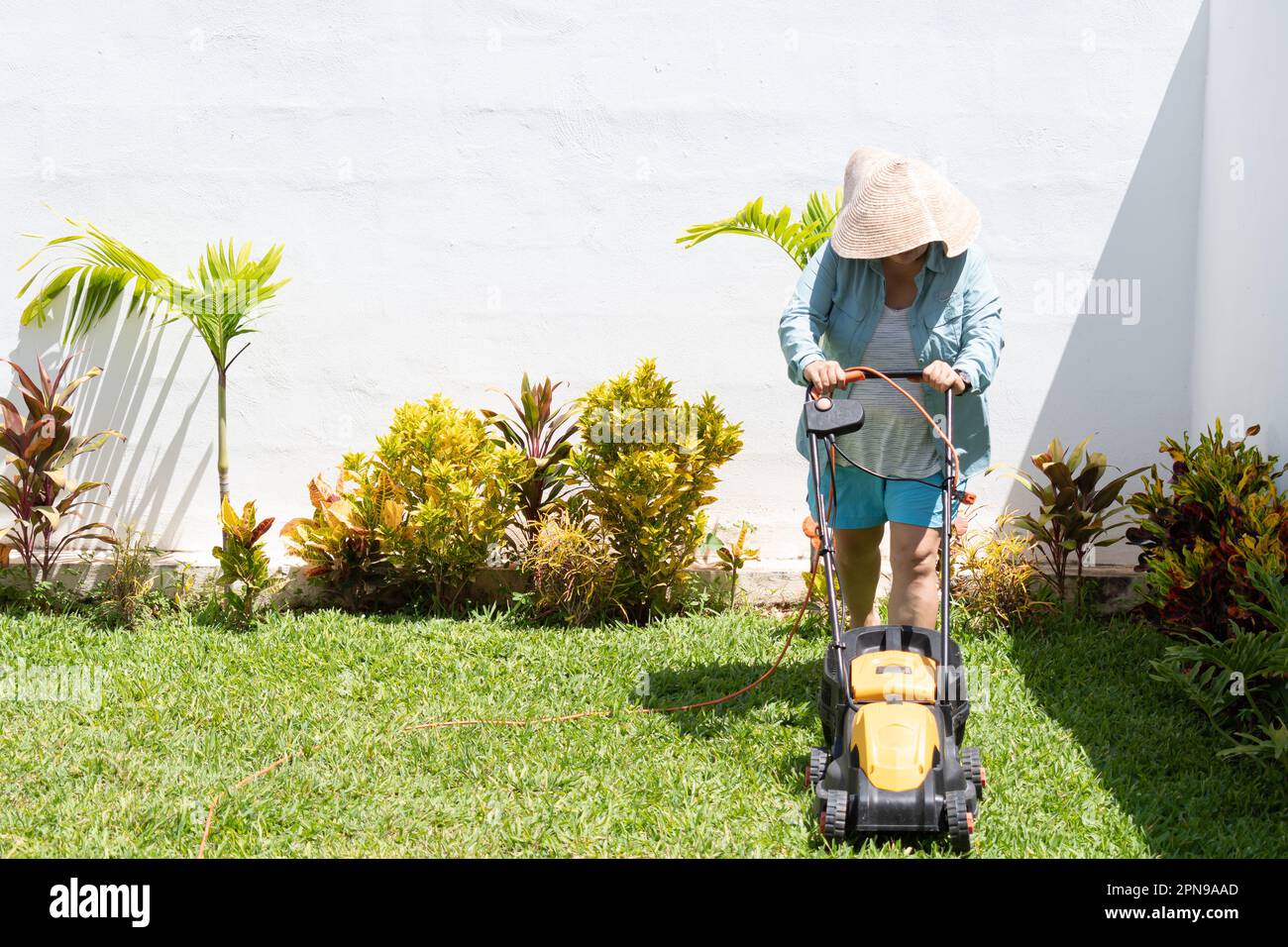 Femme portant un short bleu, une chemise à manches longues et un chapeau, tond l'herbe dans le jardin de sa maison avec une tondeuse à gazon. Banque D'Images