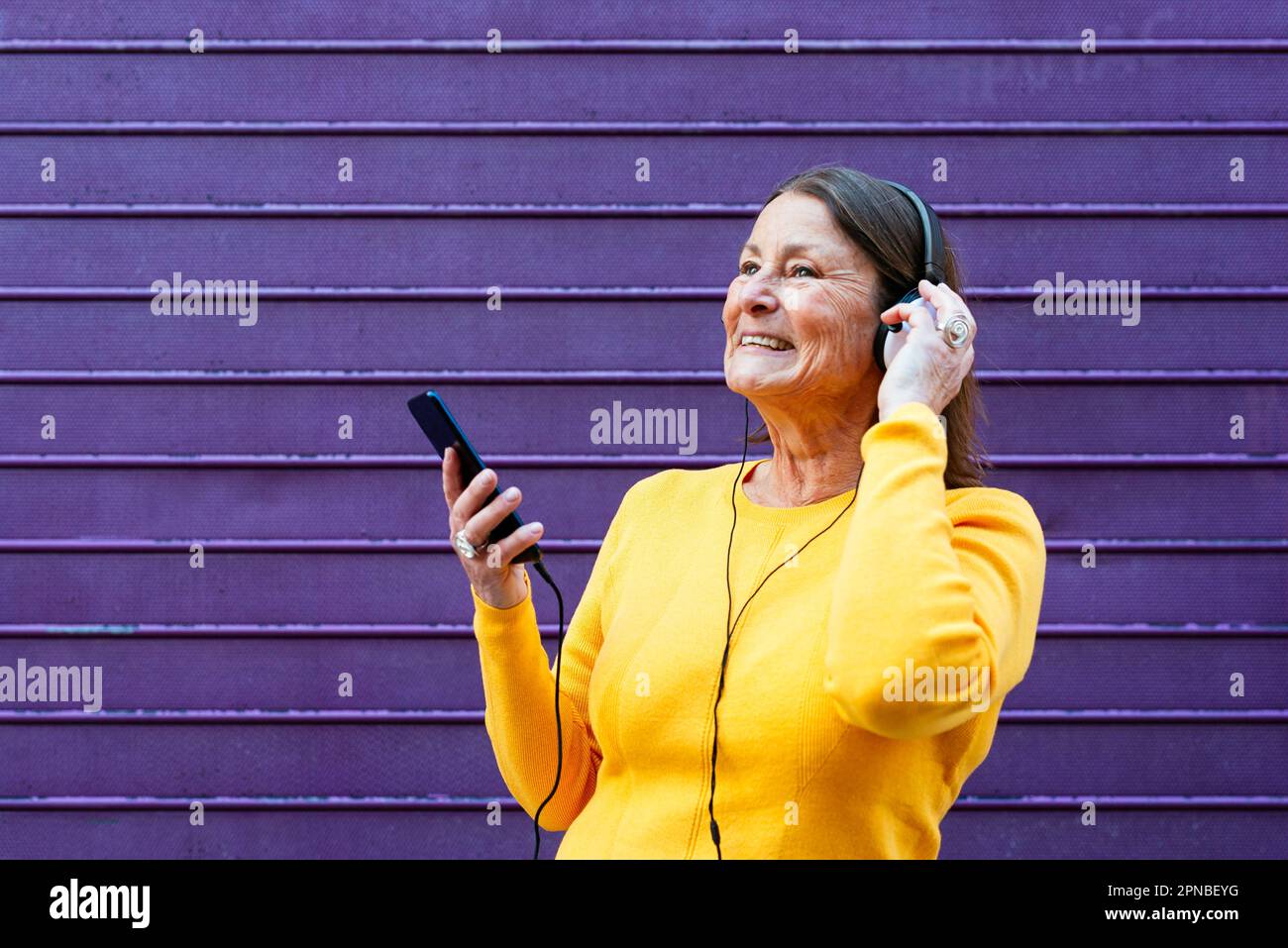 Une femme heureuse en chandail jaune qui écoute de la musique dans les écouteurs et qui navigue sur un téléphone portable tout en se tenant contre un mur violet Banque D'Images