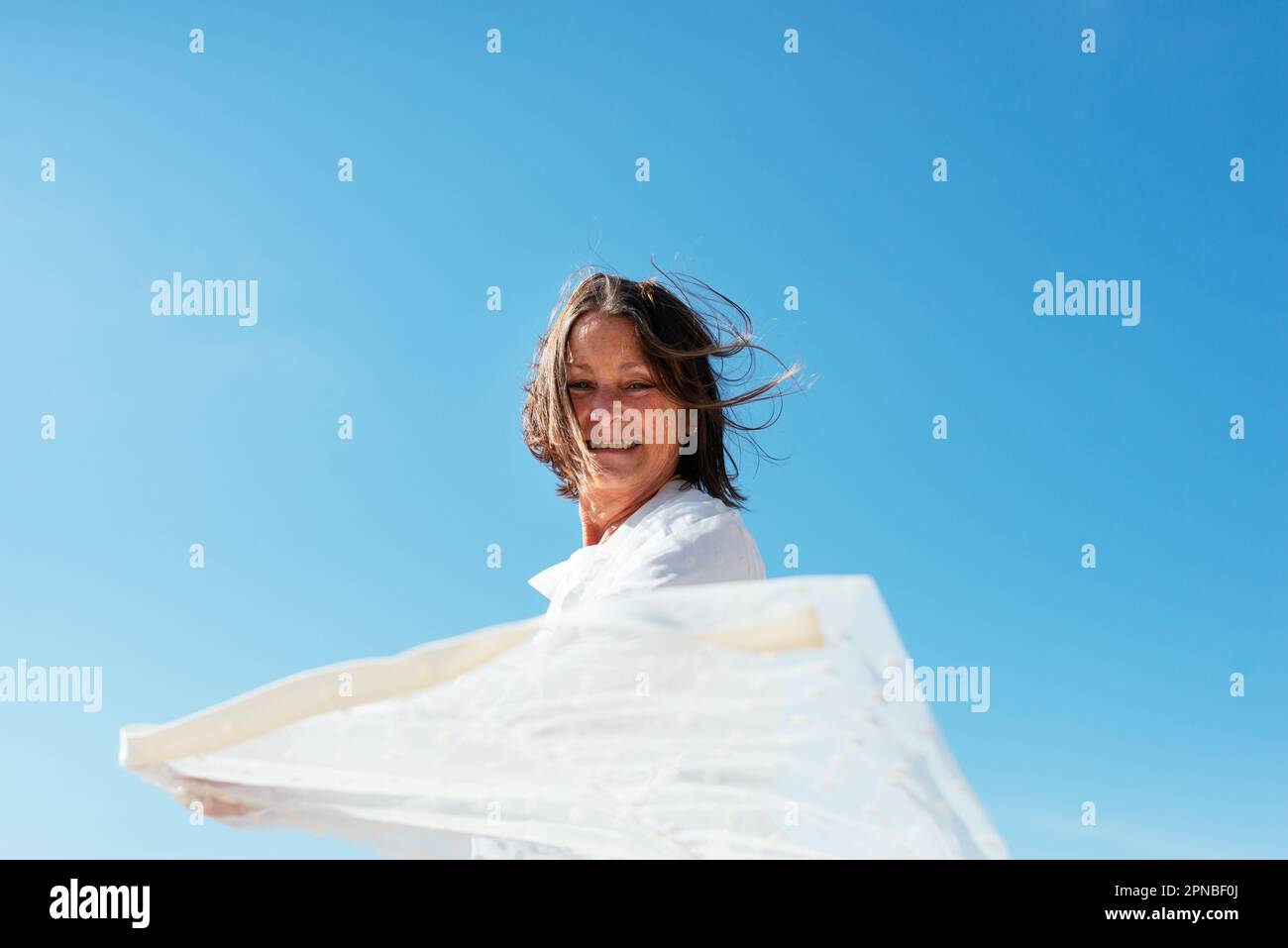 Une femme positive portant des vêtements décontractés et un manteau de plage blanc s'étirant les mains contre le ciel bleu Banque D'Images