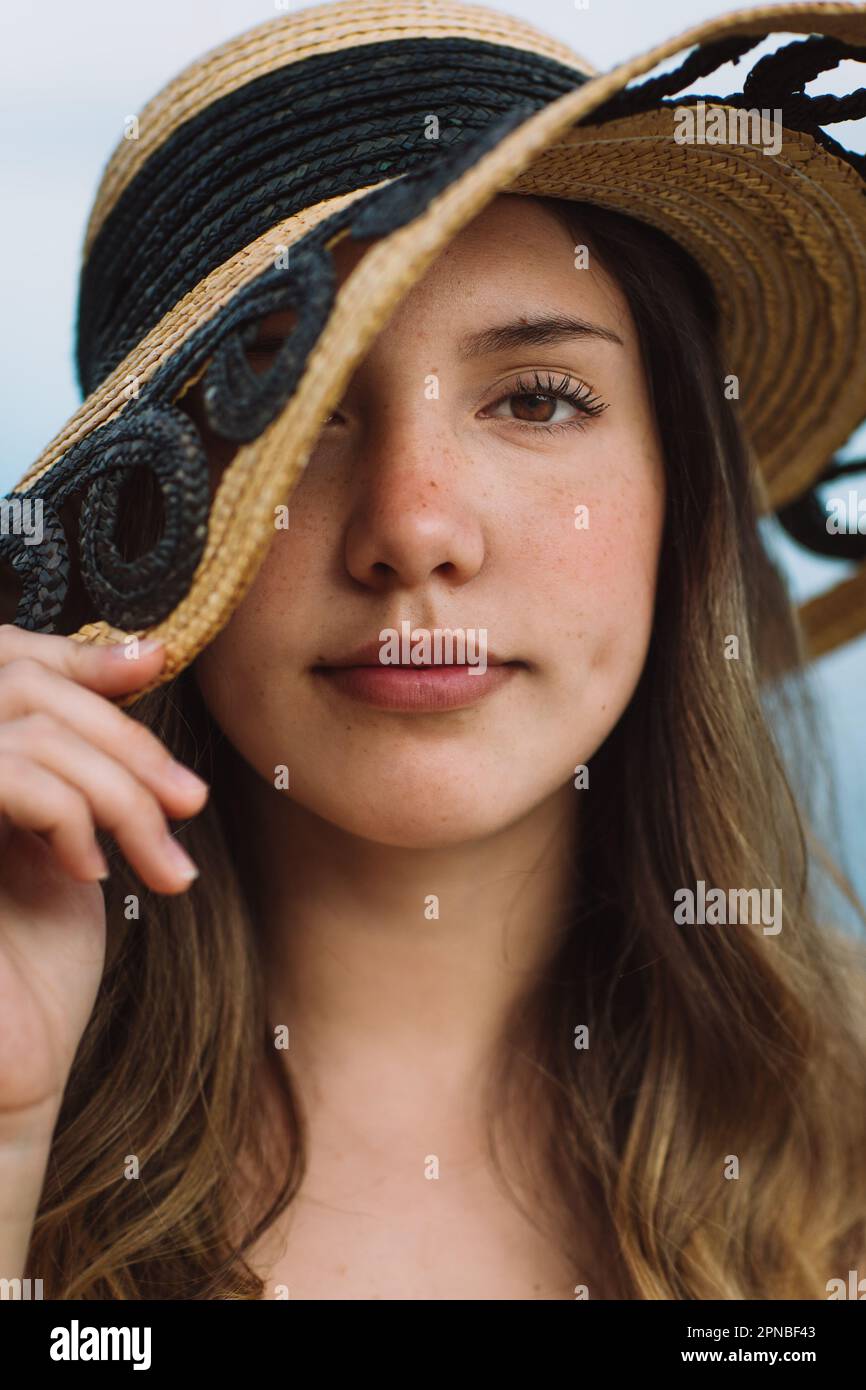 Portrait d'une femme modèle couvrant la face avec chapeau de paille et regardant l'appareil photo tout en se reposant sur les vacances d'été sur fond bleu Banque D'Images