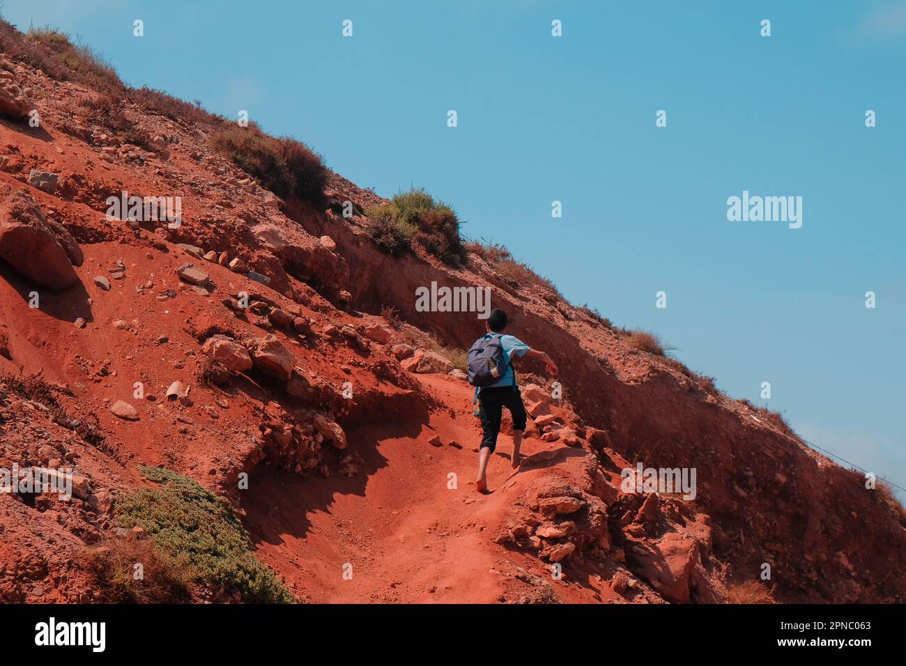 Sidi Ifni, Maroc - un garçon local avec sac à dos marche pieds nus en haut d'une colline. Jeunes Marocains randonnées à l'école. Vue de derrière. Ciel bleu. Sol en argile rouge. Banque D'Images