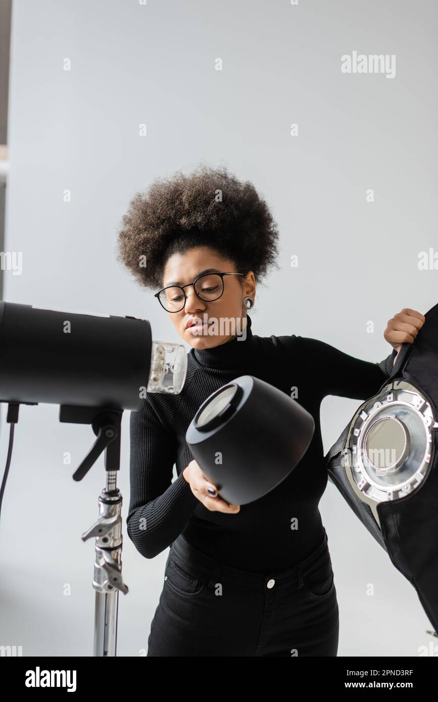 directeur de contenu afro-américain dans les lunettes tenant le réflecteur tout en assemblant la lampe stroboscopique dans le studio photo, image de stock Banque D'Images