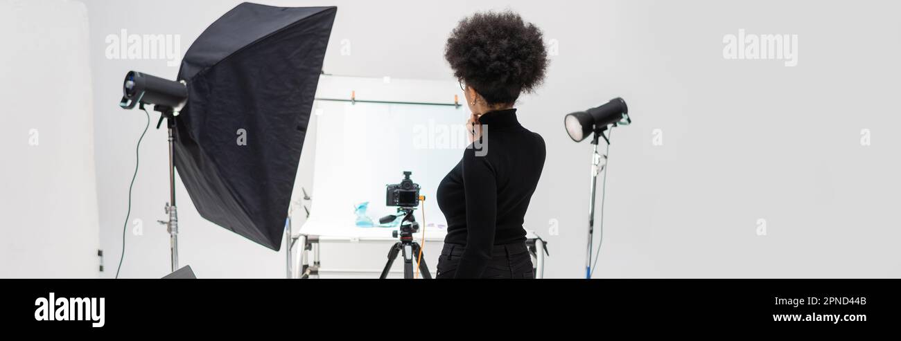 vue arrière du gestionnaire de contenu afro-américain en vêtements noirs près de l'équipement d'éclairage et de l'appareil photo numérique en studio photo, bannière, image de stock Banque D'Images
