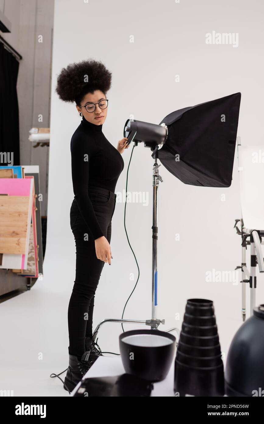 producteur de contenu afro-américain dans les lunettes et les vêtements noirs regardant l'équipement d'éclairage dans le studio photo, image de stock Banque D'Images