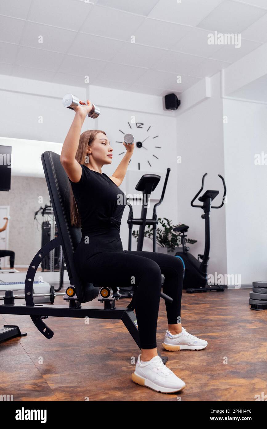 La jeune femme blondy sportive en noir fait de la dumbbell presse sur le banc universel incliné à la salle de gym ou au studio de fitness. Pompage des muscles de b Banque D'Images