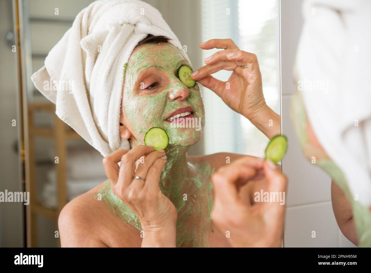 Mature belle femme appliquant le masque facial vert maison DIY sur la peau. Regarder dans le miroir dans la salle de bain, s'amuser. Enveloppé dans une serviette. Soins de la peau Banque D'Images