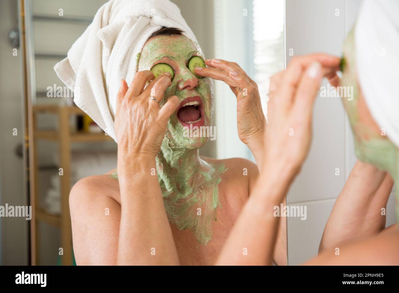 Mature belle femme appliquant le masque facial vert maison DIY sur la peau. Regarder dans le miroir dans la salle de bain, s'amuser. Enveloppé dans une serviette. Soins de la peau Banque D'Images