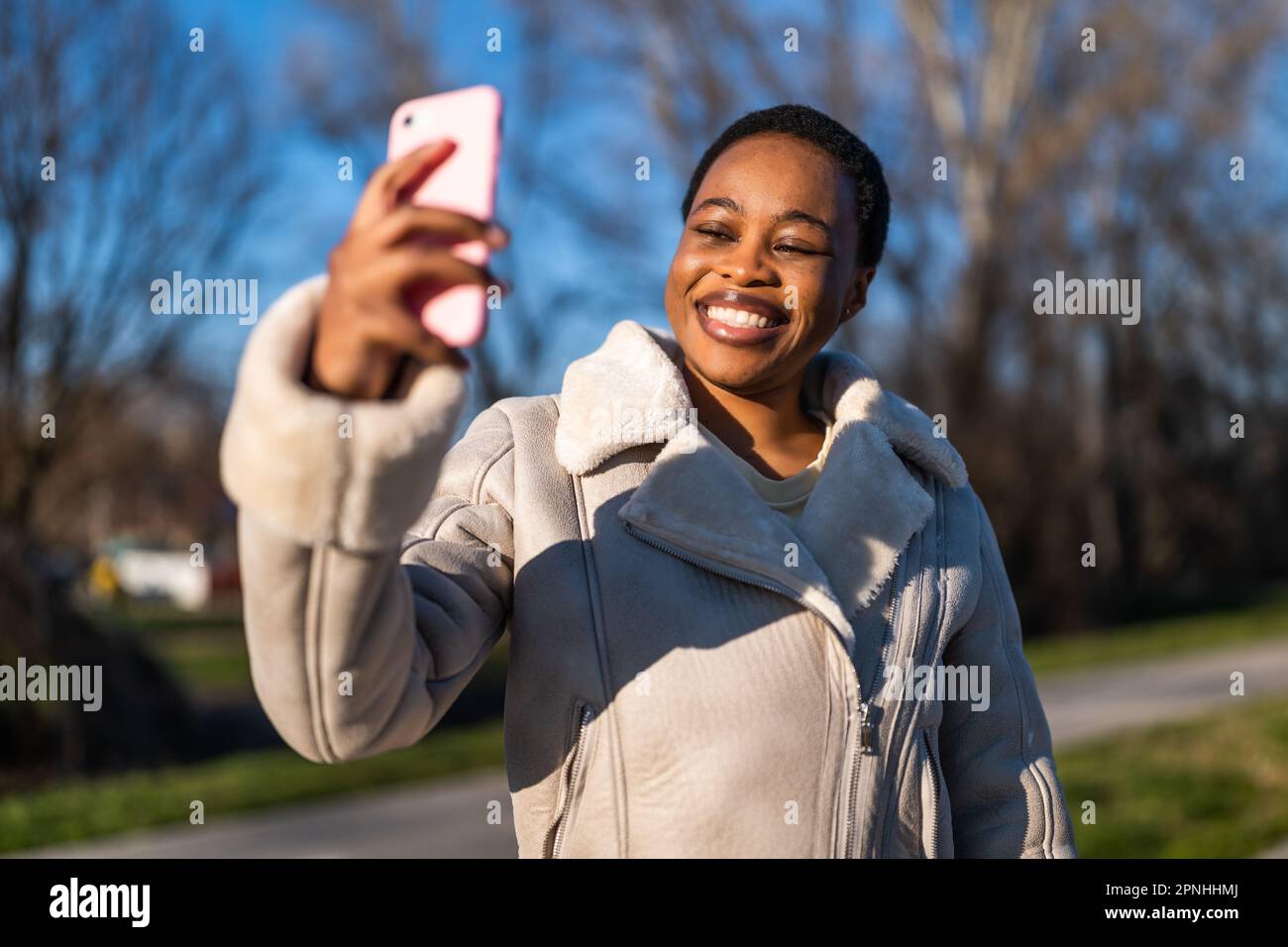 Portrait extérieur d'une femme noire heureuse le jour ensoleillé. Elle est assise sur le banc dans la rue et prend le selfie. Banque D'Images