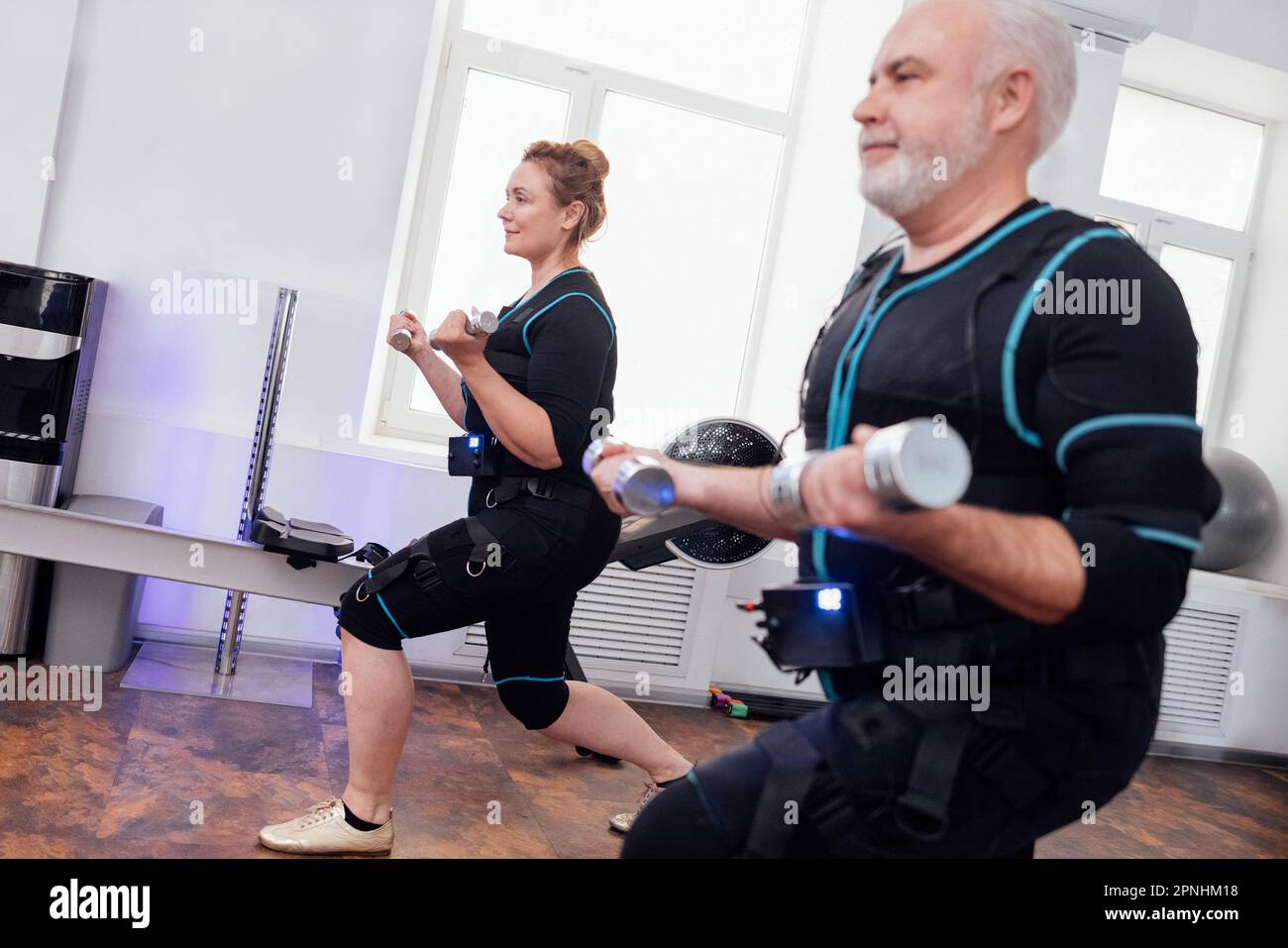 Couple de personnes âgées souriant dans des combinaisons ems faisant des exercices physiques avec des haltères dans la salle de gym. Homme à poil gris et femme à poil dur s'entraîner avec un t personnel Banque D'Images