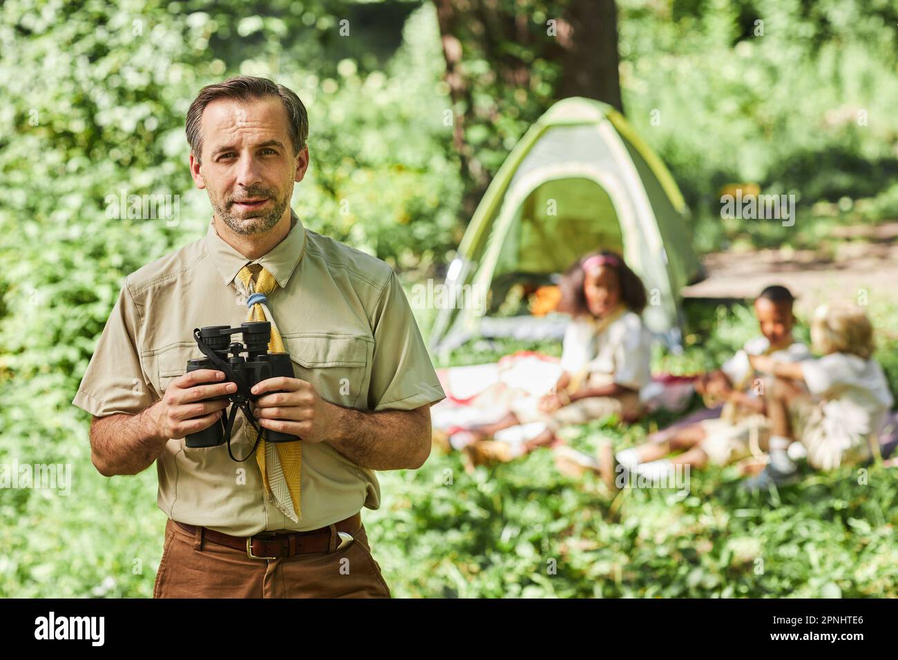 Montez le portrait d'un homme mûr en tant que chef de scout tenant des jumelles et regardant l'appareil-photo pendant le camping avec le groupe scolaire, copier l'espace Banque D'Images