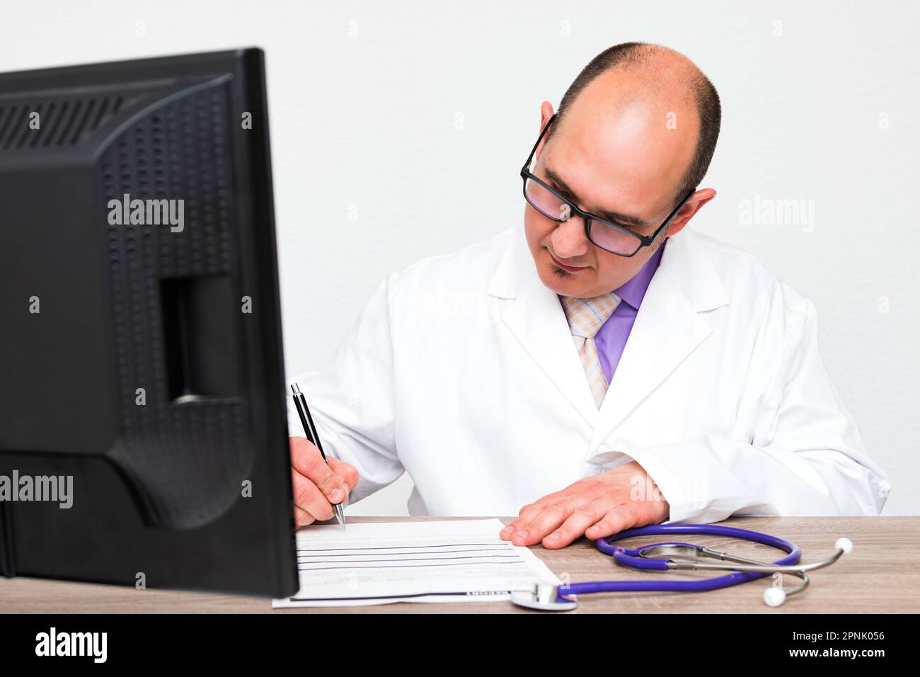 Un médecin caucasien chauve avec des lunettes portant un manteau blanc signant un rapport de santé sur sa table en bois de bureau. Banque D'Images