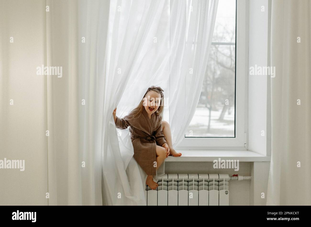 Portrait d'une petite fille dans une robe de chambre brune, qui est assise sur le rebord de la fenêtre. Banque D'Images