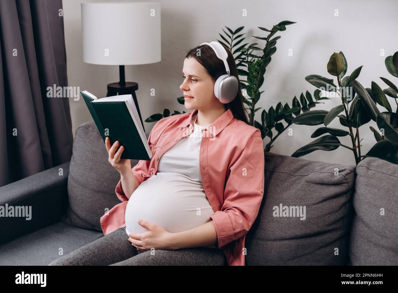 Belle jeune femme enceinte caucasienne écoutez de la musique avec un casque sans fil et un livre de lecture pour raconter une histoire à un enfant à naître. Beau futur calme Banque D'Images