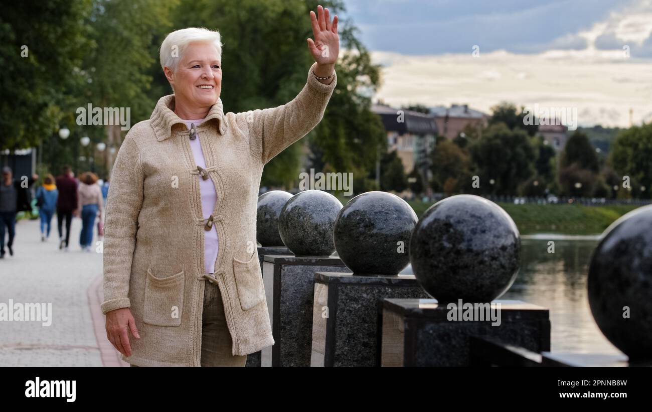 Femme aux cheveux gris retraite belle femme positive dans des vêtements chauds se détendre à l'extérieur par temps frais, en regardant loin bonjour ou au revoir, souriant Banque D'Images