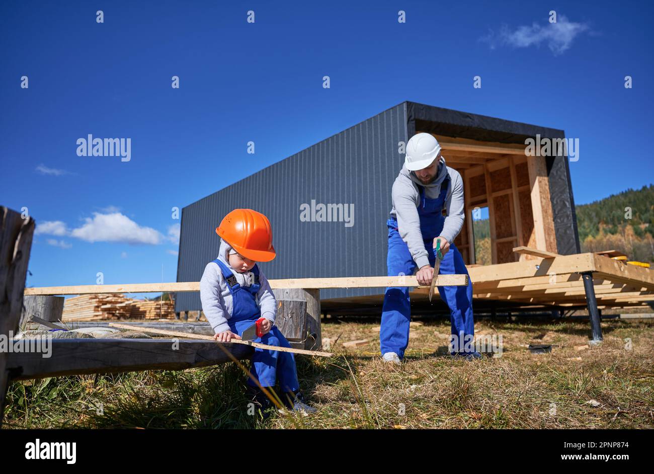 Père avec enfant fils bâtiment en bois maison de cadre. Garçon aidant son papa, en utilisant la scie à main pour couper des planches sur le chantier, portant un casque, des combinaisons bleues le jour ensoleillé. Menuiserie et concept de famille. Banque D'Images