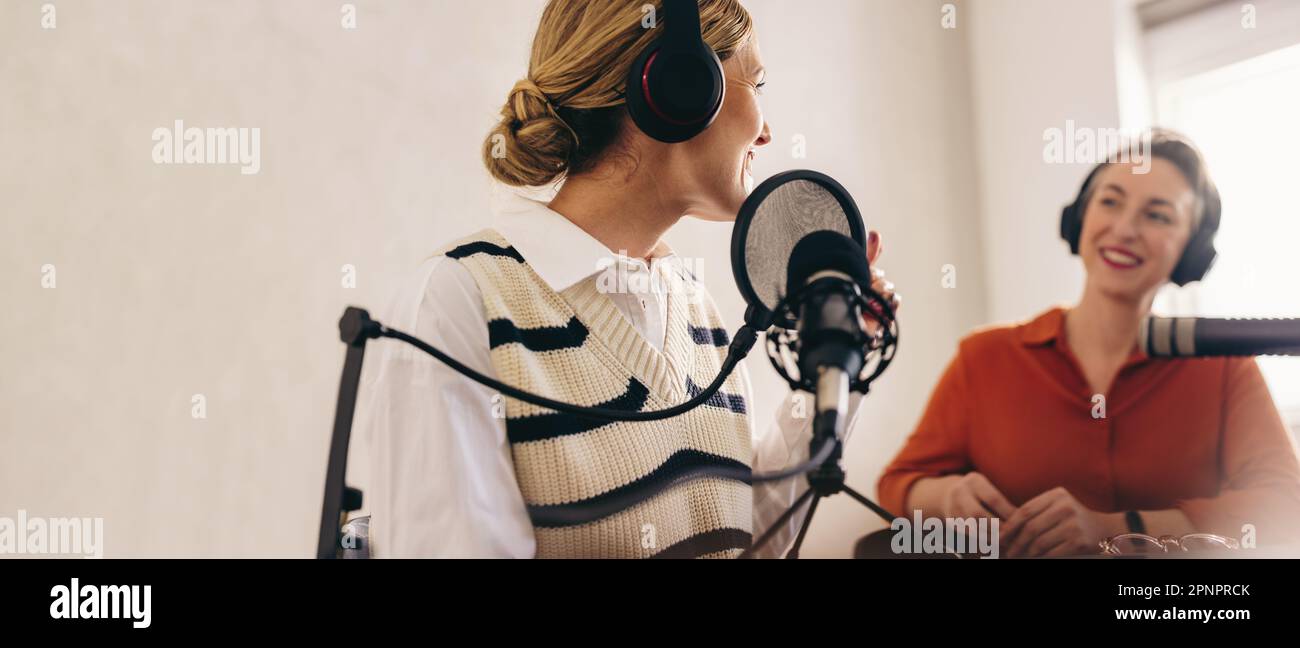 Une heureuse hôtesse de radio interrogeant une cliente sur son spectacle dans son studio à domicile. Deux femmes gaies ayant un bon moment tout en enregistrant une émission audio en direct sur Banque D'Images