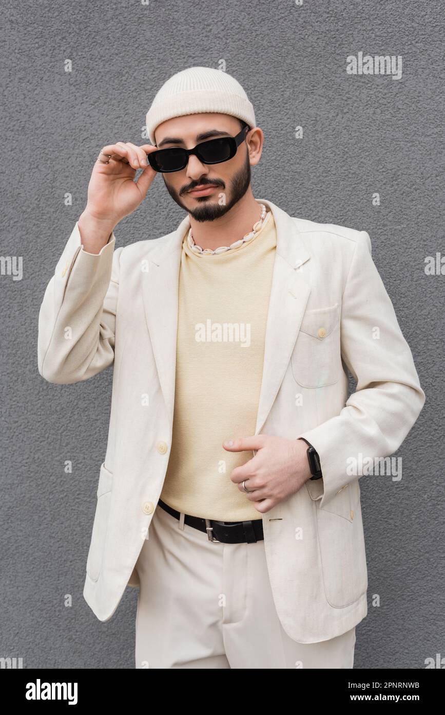Homme homosexuel tendance en costume beige touchant lunettes de soleil à l'extérieur, image de stock Banque D'Images