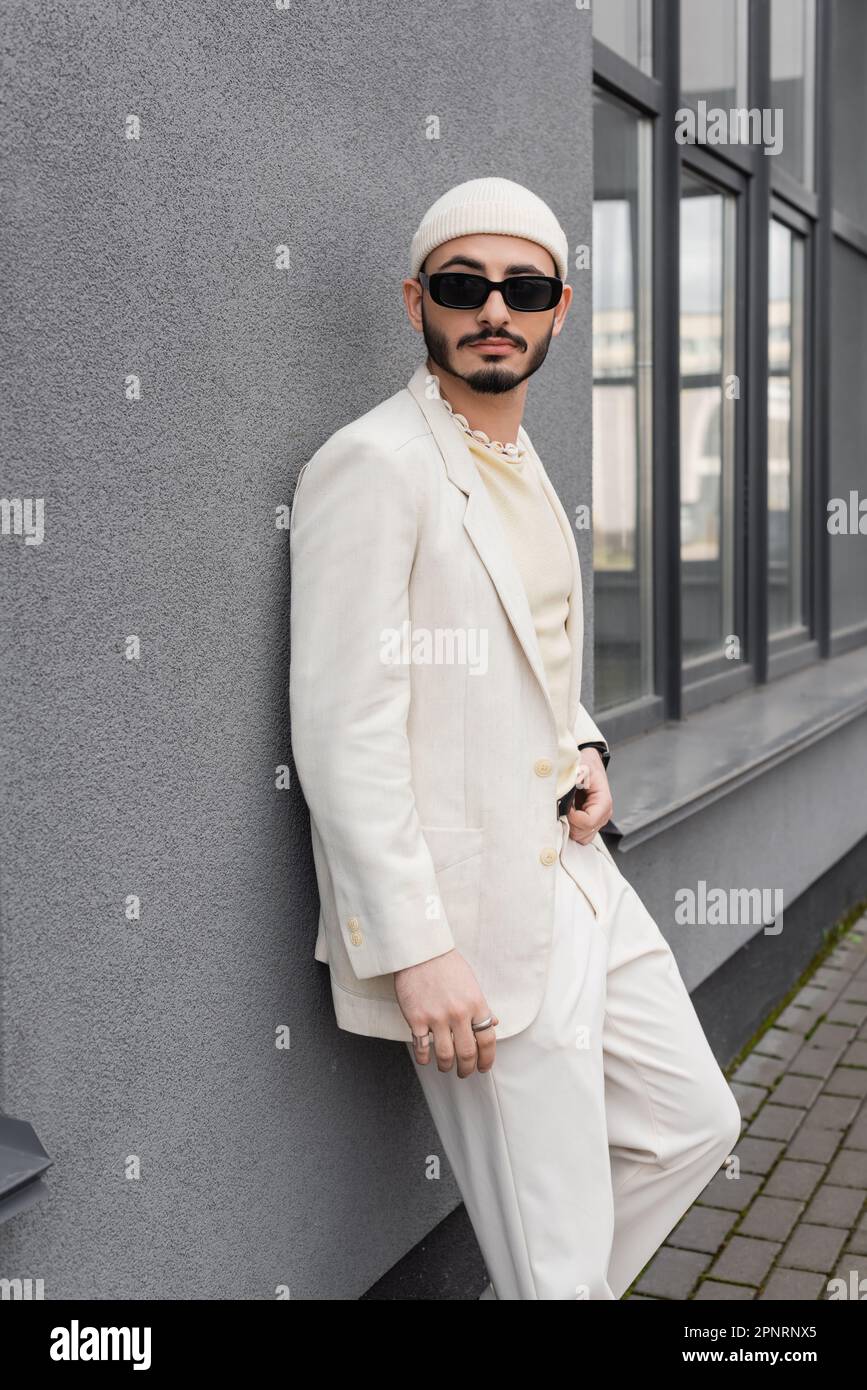 Homme homosexuel élégant en lunettes de soleil et costume debout près de la façade du bâtiment à l'extérieur, image de stock Banque D'Images