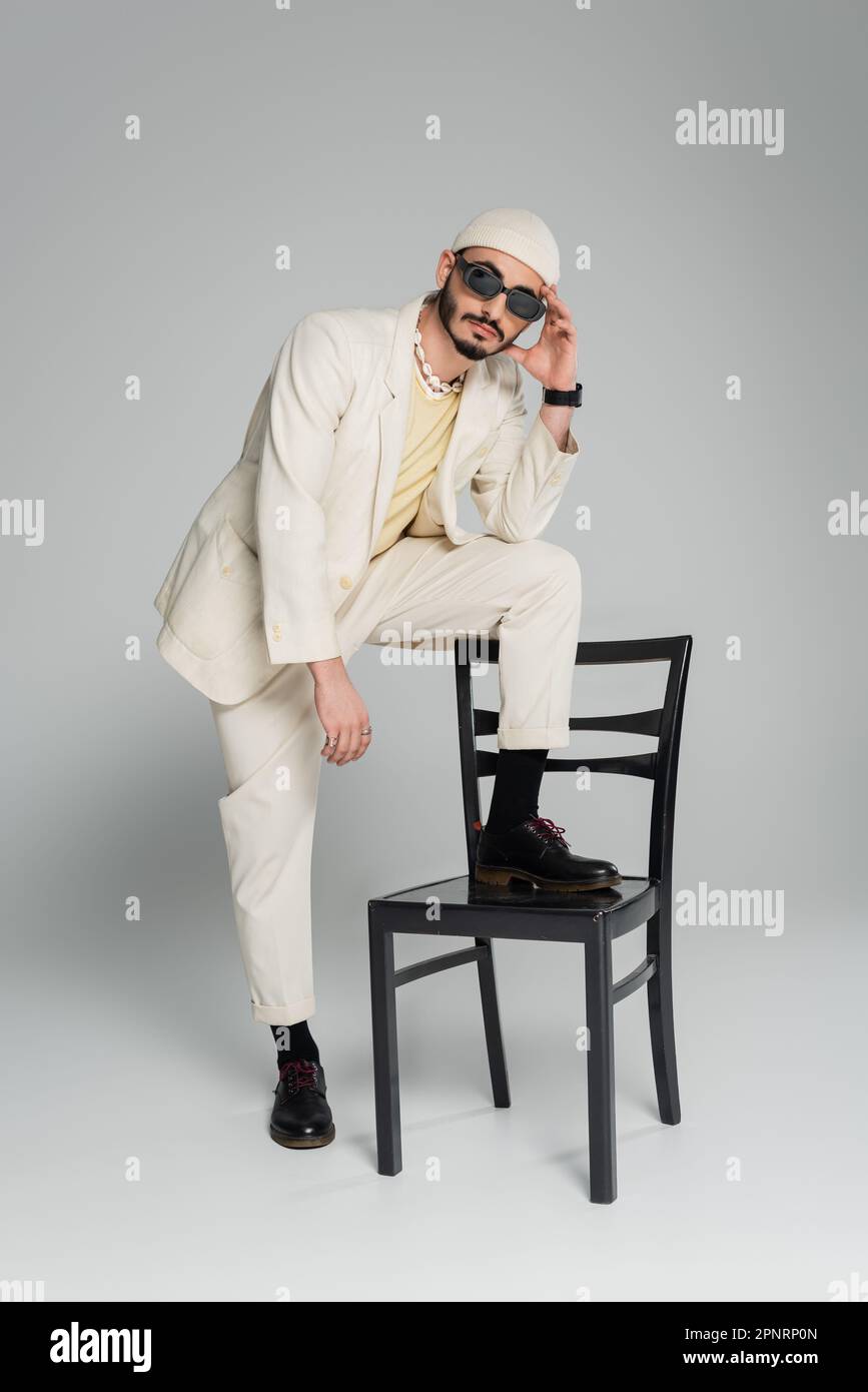Homme homosexuel à la mode en costume et chapeau posant près de la chaise sur fond gris, image de stock Banque D'Images