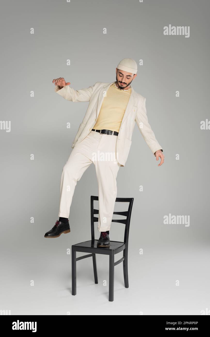 Homme homosexuel à la mode en costume et chapeau beige debout sur la chaise sur fond gris, image de stock Banque D'Images