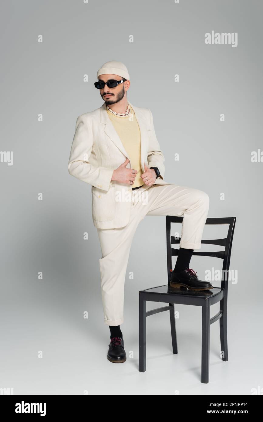 Homme homosexuel à la mode dans des lunettes de soleil posant près de la chaise sur fond gris, image de stock Banque D'Images