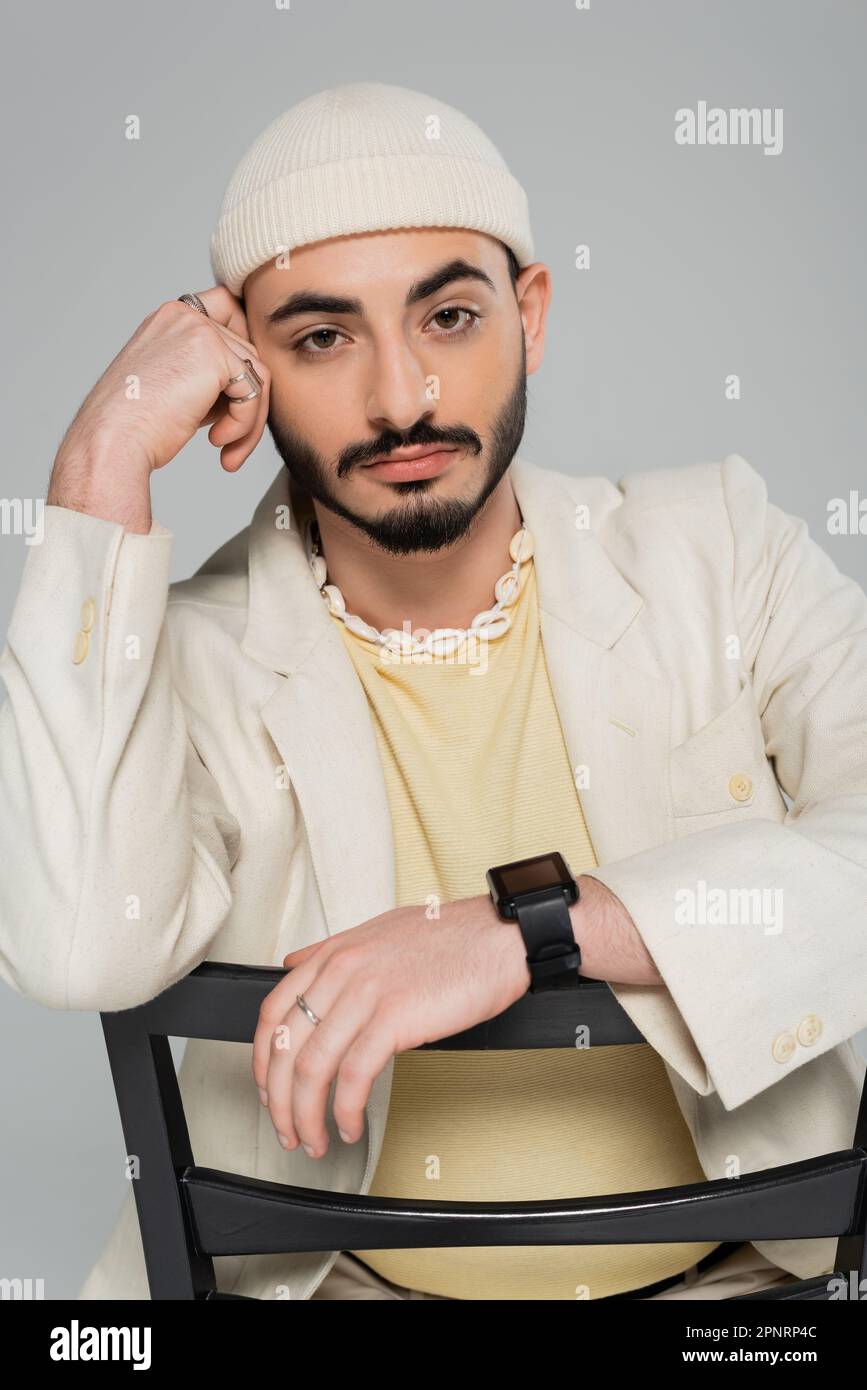Portrait de l'homme homosexuel branché dans un chapeau et une veste assis sur une chaise isolée sur une image grise Banque D'Images