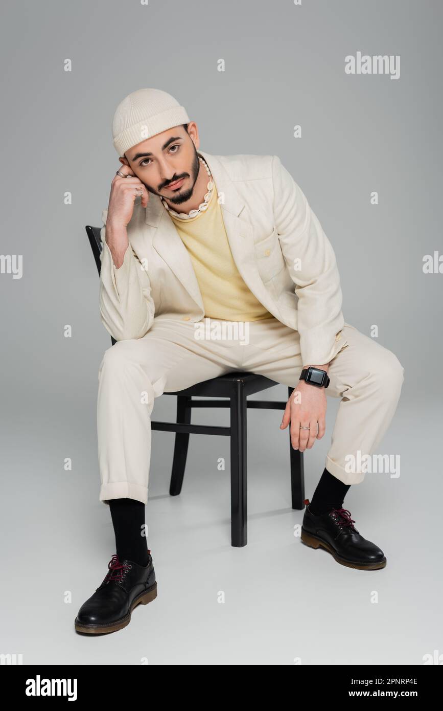 Pleine longueur d'homme homosexuel élégant en costume et chapeau regardant l'appareil photo sur fond gris, image de stock Banque D'Images