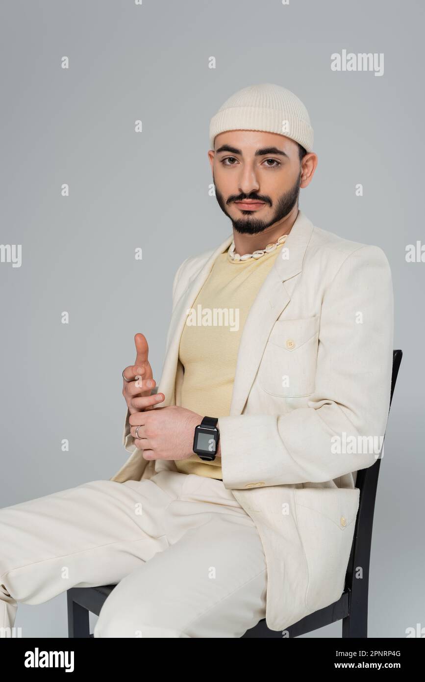 Homme homosexuel barbu en chapeau et costume assis sur une chaise isolée sur image grise Banque D'Images