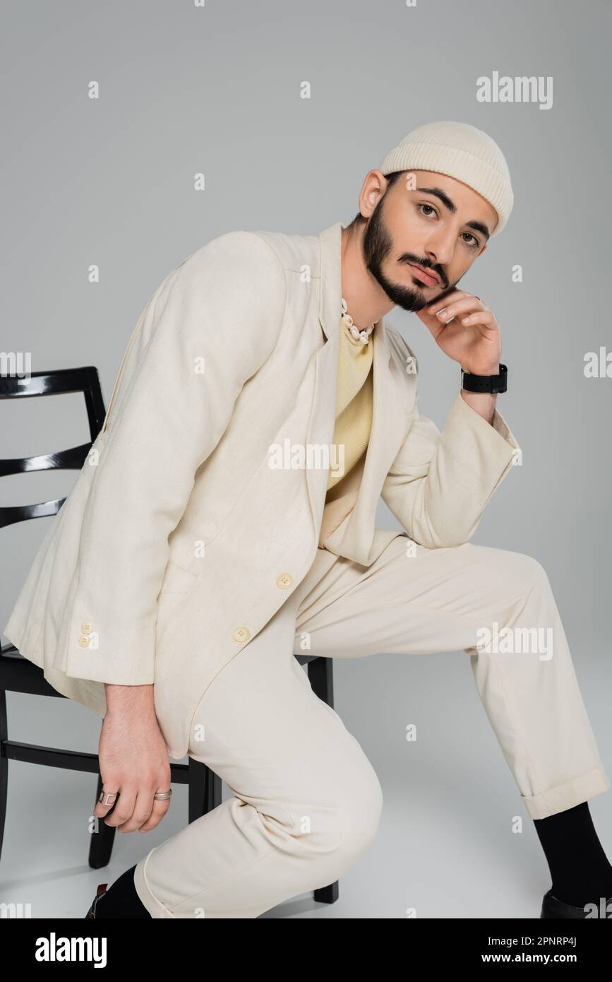 Homme homosexuel tendance en chapeau et costume beige posant sur la chaise sur fond gris, image de stock Banque D'Images