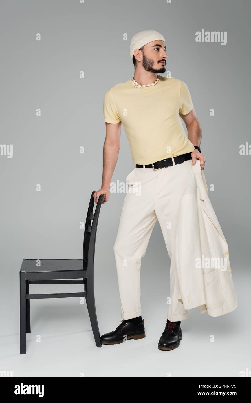 Pleine longueur de l'homme homosexuel bien habillé tenant la veste près de la chaise sur fond gris, image de stock Banque D'Images