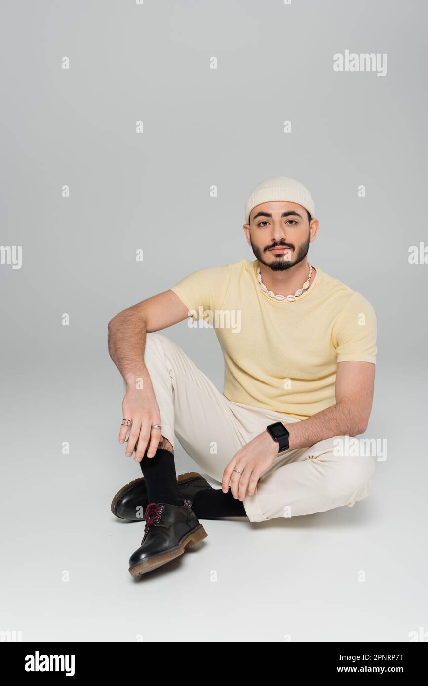 Homme homosexuel barbu en chapeau regardant la caméra tout en étant assis sur fond gris, image de stock Banque D'Images