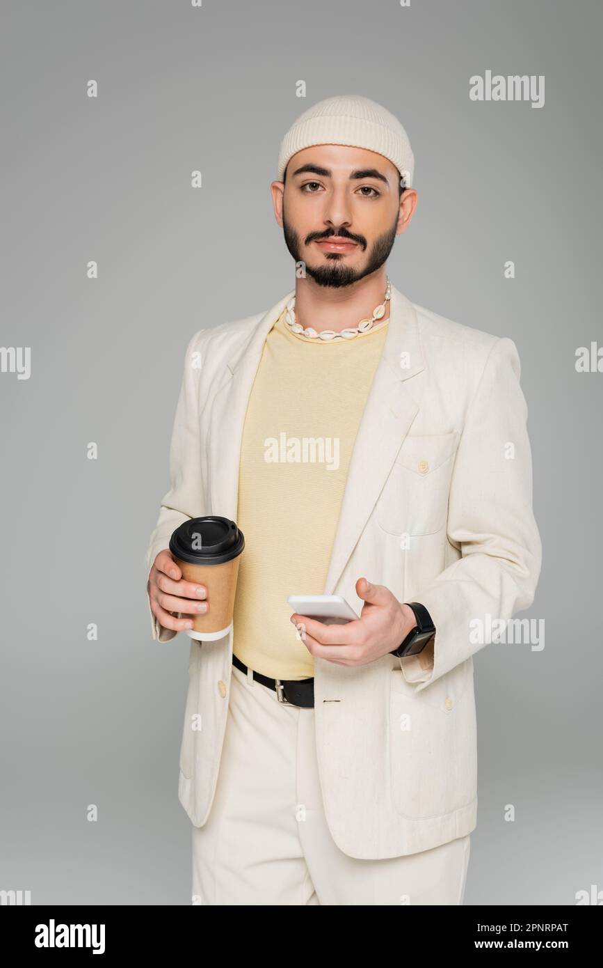 Homme homosexuel élégant en costume tenant smartphone et café pour être isolé sur gris, image de stock Banque D'Images