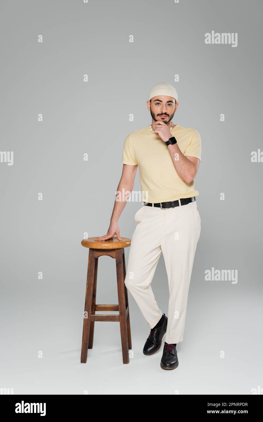 Homme homosexuel élégant en chapeau et montre intelligente debout près de la chaise sur fond gris, image de stock Banque D'Images