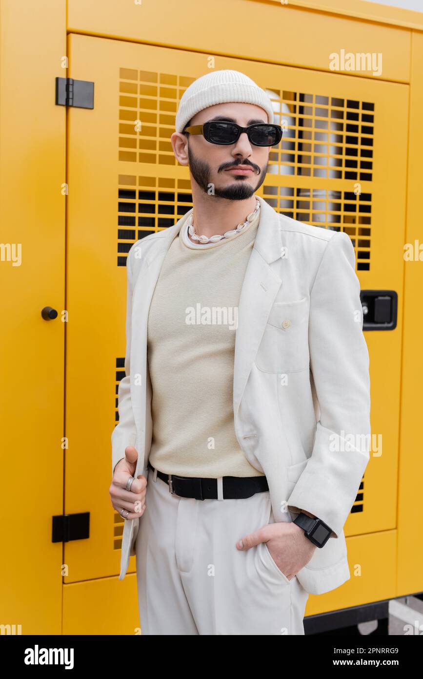 Homme homosexuel élégant en costume beige et lunettes de soleil debout sur la rue urbaine, image de stock Banque D'Images
