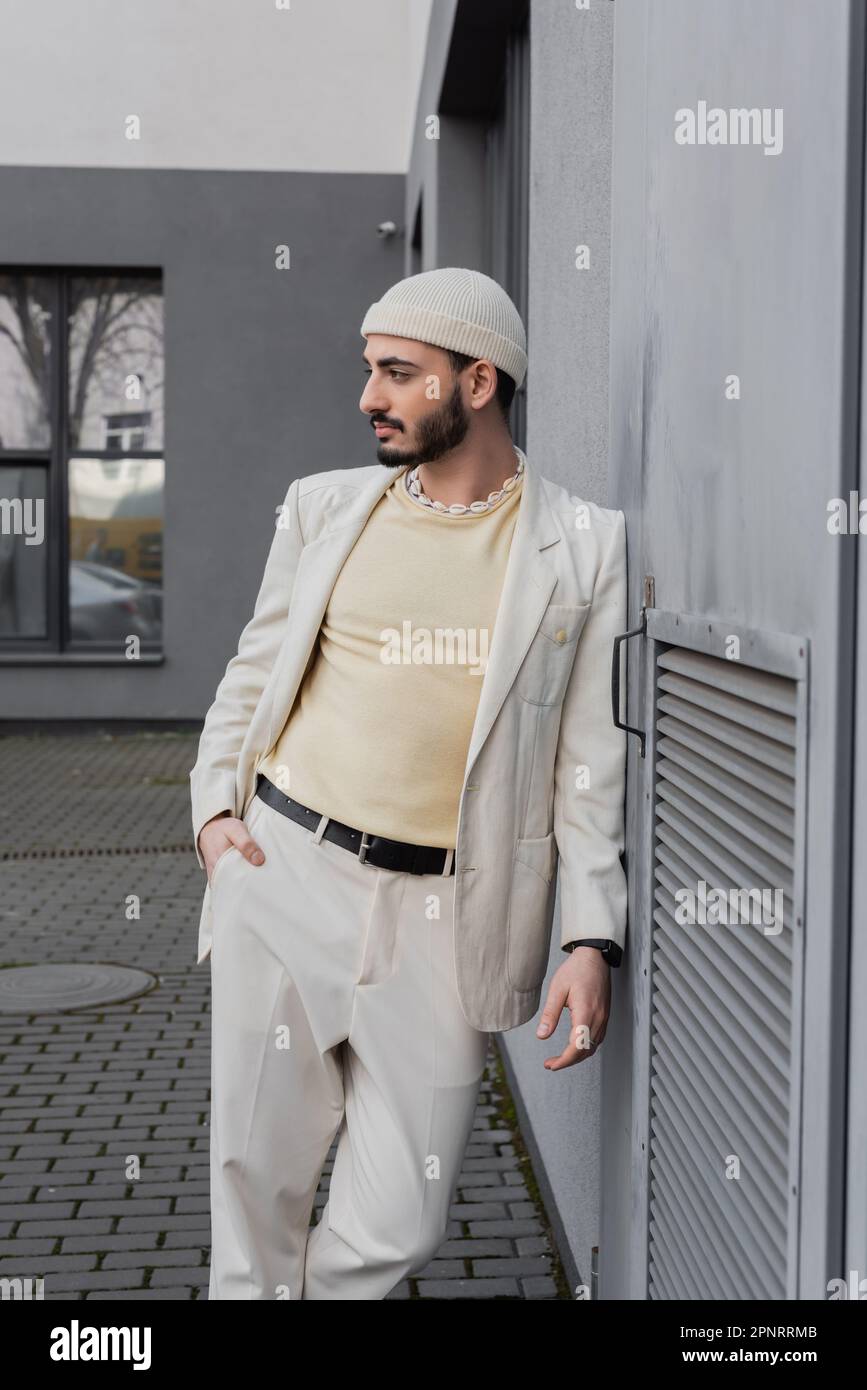 Homme homosexuel élégant en chapeau beige posant près du mur de bâtiment à l'extérieur, image de stock Banque D'Images