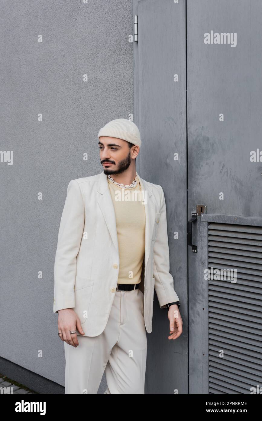 Homme homosexuel tendance en chapeau et costume regardant loin de bâtiment sur la rue urbaine, image de stock Banque D'Images