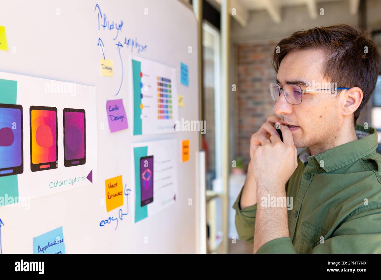 Un designer caucasien parlant sur un smartphone, regardant des maquettes et des notes sur un tableau blanc en studio Banque D'Images