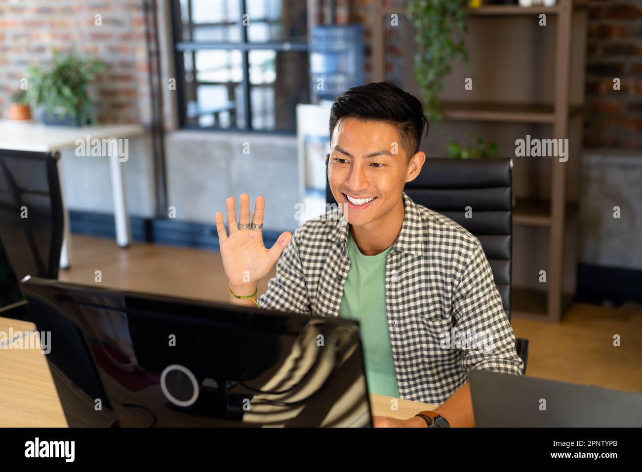 Souriant homme asiatique créatif à l'ordinateur faire des appels vidéo, agitant dans un bureau décontracté Banque D'Images