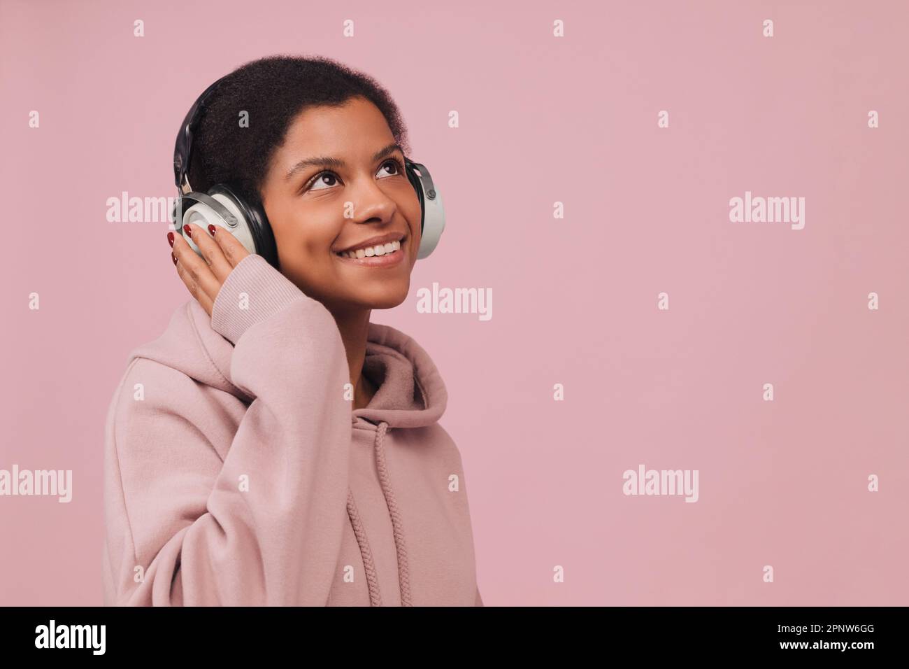 Photo en gros plan d'une jeune femme noire profitant de la musique des écouteurs. Jolie fille gaie avec écouteurs blancs sur fond rose Banque D'Images