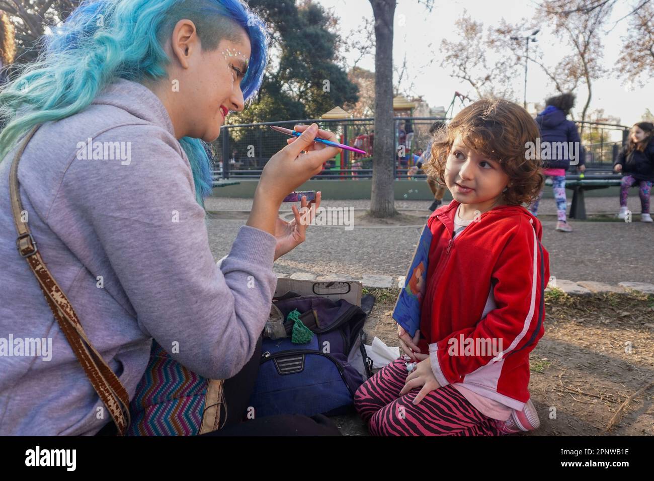 Magalí Murúa se prépare à mettre des paillettes sur la joue de Lola Ferreyra Puchatt, 4 ans, dans un parc à Buenos Aires, en Argentine, sur 22 juillet 2022. Depuis qu'elle a perdu son emploi en 2020, Murúa a peint des visages dans des manifestations, des parcs et des événements. Elle price ses services sur une échelle mobile, de sorte qu'ils sont abordables pour tous. (Lucila Pellettieri/Global Press Journal) Banque D'Images